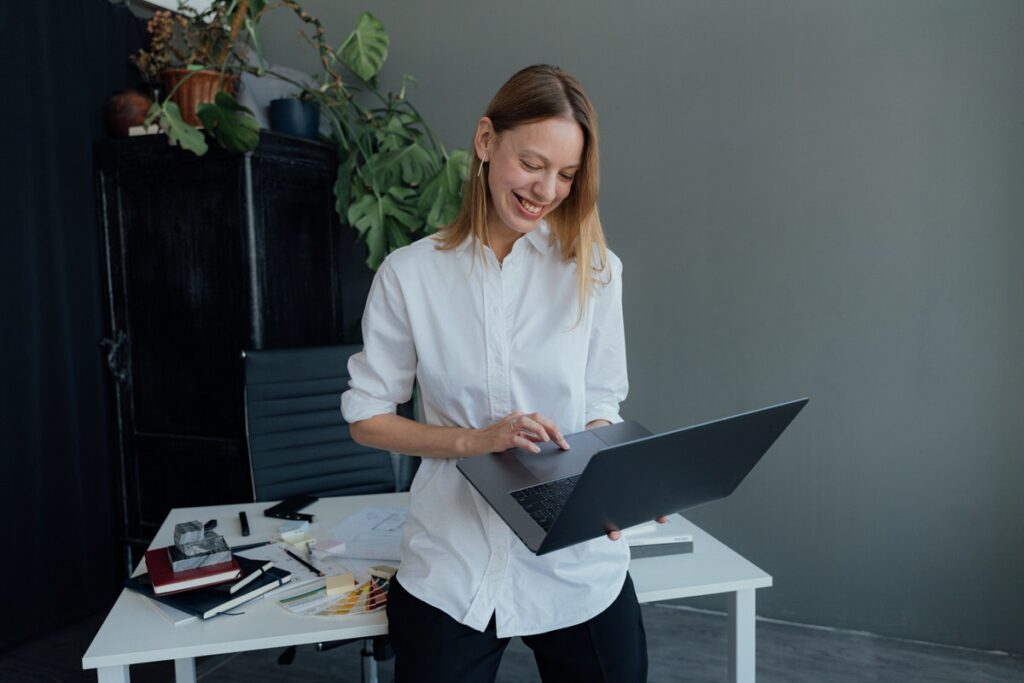 Woman standing in front of a desk while holing an open laptop. 
