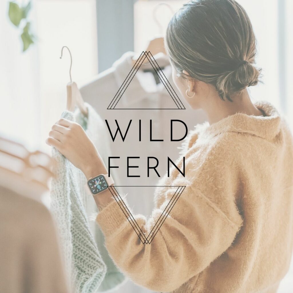 Woman holding up two different sweaters in a store with a logo over the middle of the image that reads "Wild Fern".