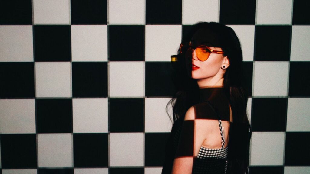woman in sunglasses standing against a black and white checkered board wall with shadows over her.