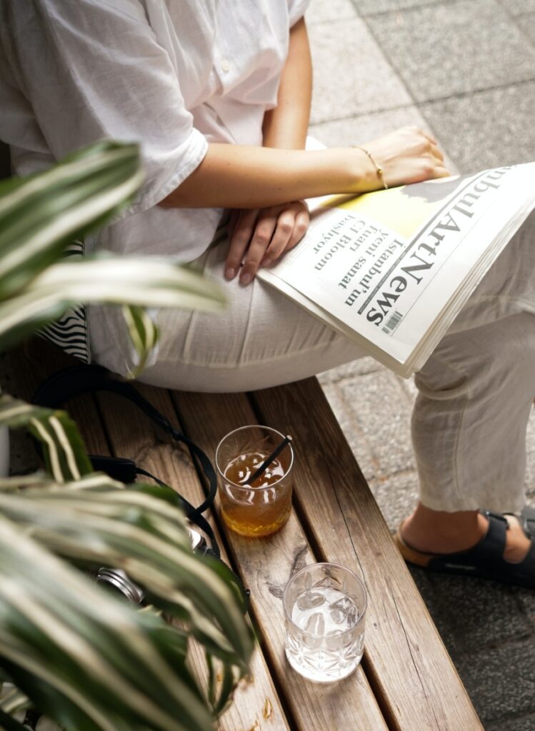 Person sitting on a bench next to two drinks while they read a newspaper.