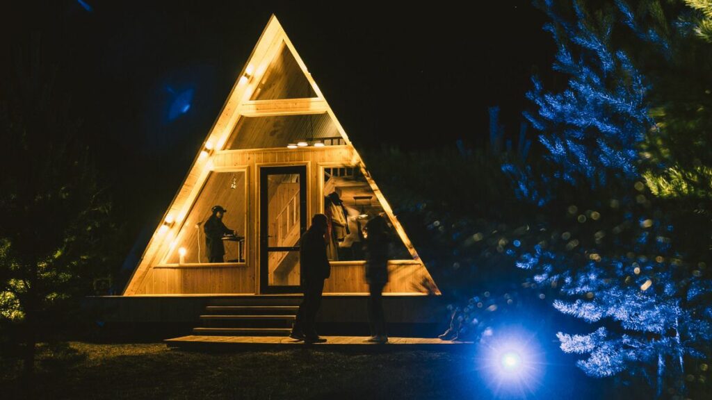A frame shaped house lit up against the night sky with people walking up the stairs into it 