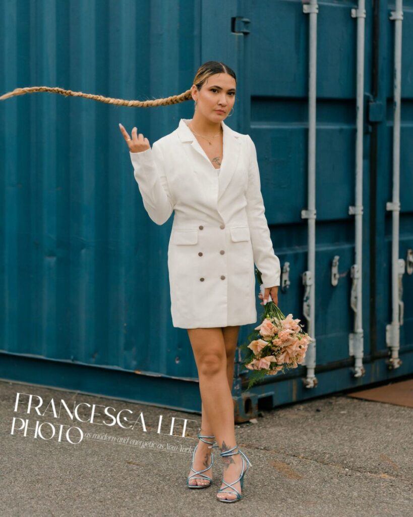 Person standing in front of a blue shipping container flipping her braid over her should with one hand while holding a bouquet with the other. In the bottom left hand corner there is a white logo that says "Francesca Lee Photo. As modern and energetic as New York."