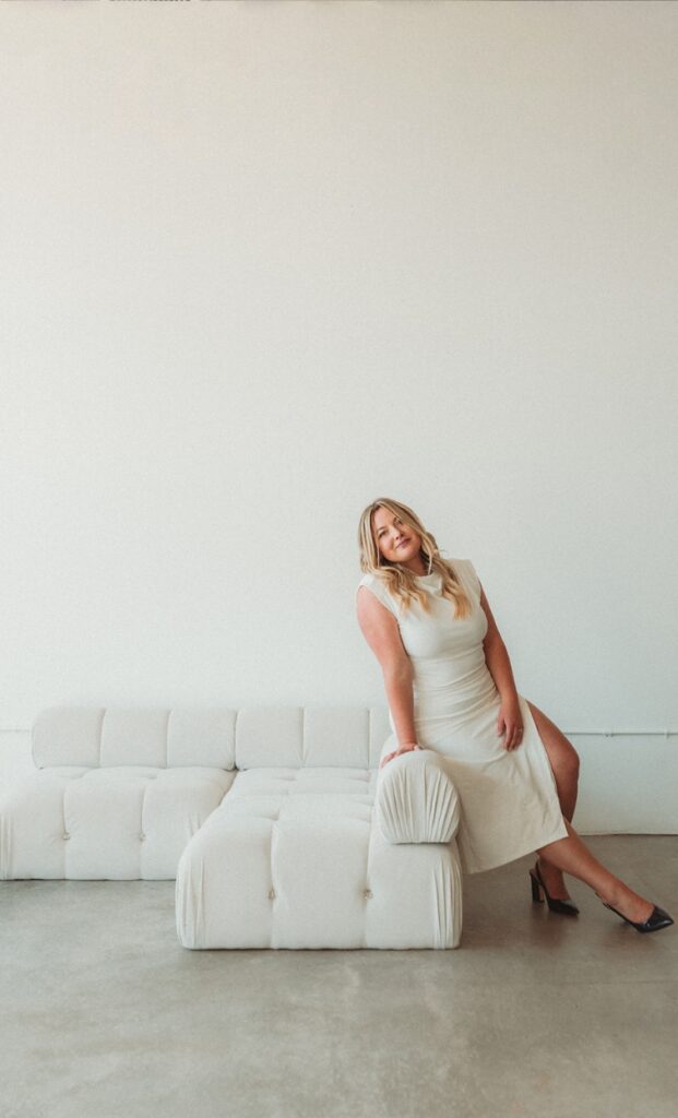 Woman in a white dress leaning against a couch.