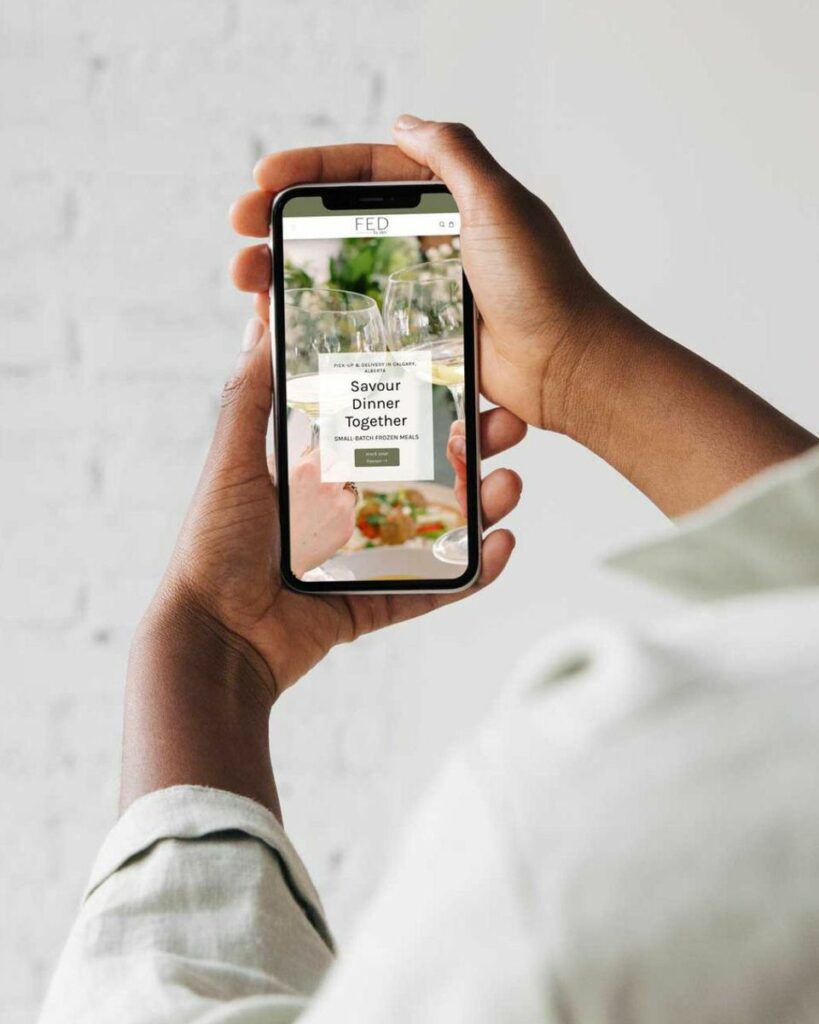 A person's hand holding a smartphone displaying the FED by Alex website with the text 'Savour Dinner Together' on a webpage showcasing small batch frozen meals