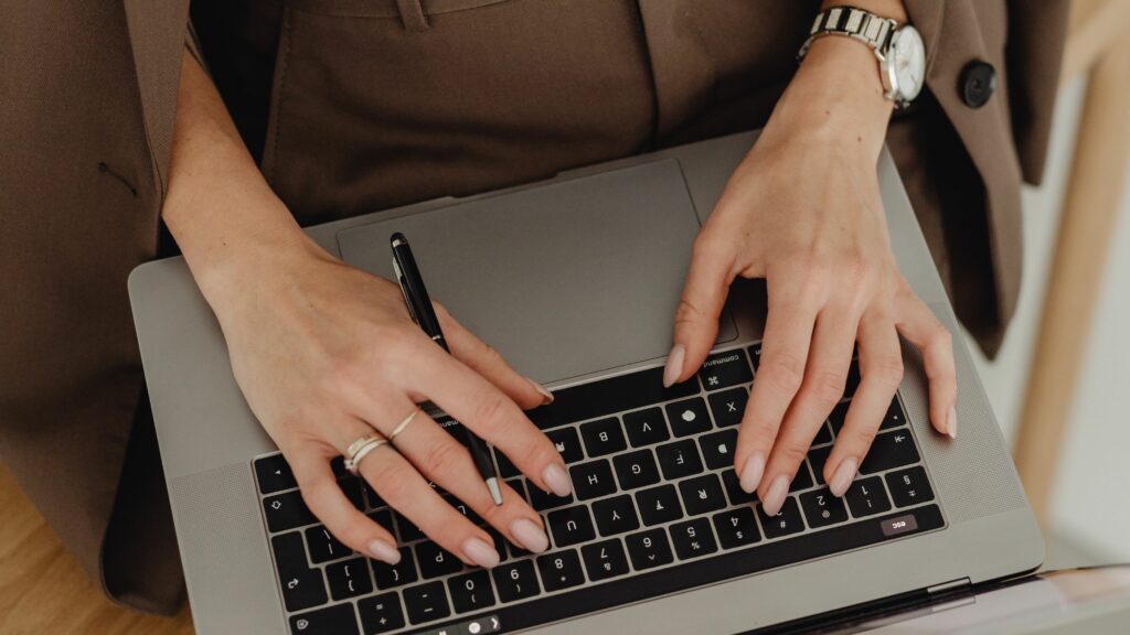 Close-up of a person's hands typing on a modern laptop keyboard, wearing a beige dress and a wristwatch.
