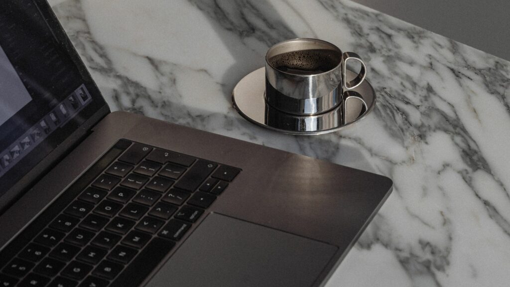 A laptop with code on the screen placed on a marble surface next to a metal cup of black coffee.