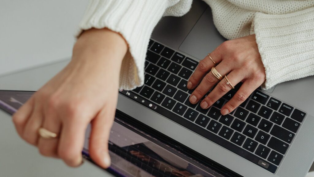 Over-the-shoulder view of a person working on a laptop, hands on the keyboard, wearing a white sweater with gold rings.