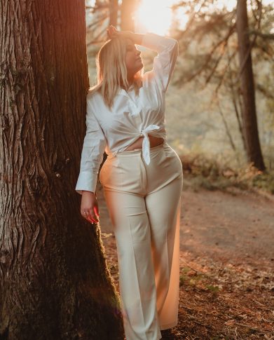 Woman leaning up against a tree with one hand on her head at sunset.